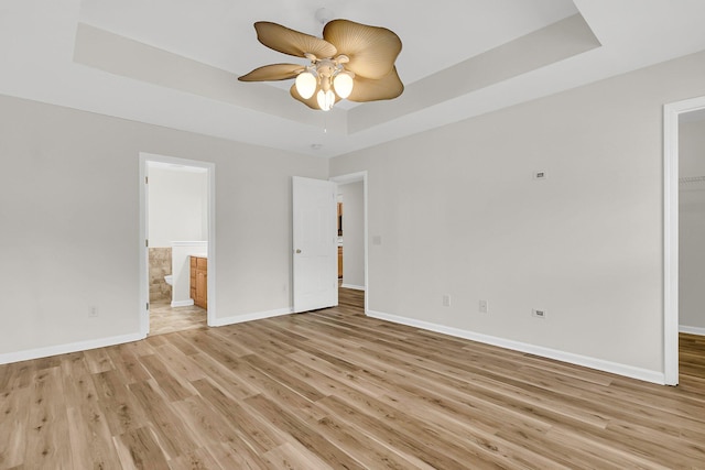 unfurnished bedroom featuring connected bathroom, baseboards, a walk in closet, light wood finished floors, and a raised ceiling