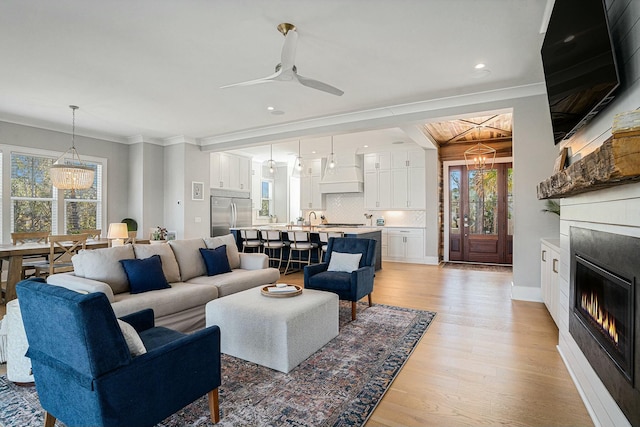 living area featuring light wood-style flooring, ceiling fan with notable chandelier, a glass covered fireplace, crown molding, and baseboards