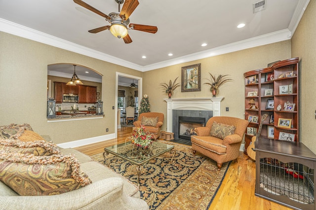 living room featuring light wood-type flooring, ceiling fan, ornamental molding, and a tiled fireplace