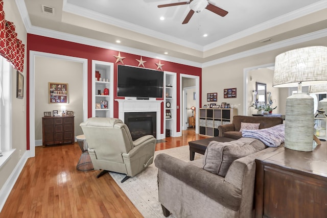 living room with ceiling fan, a raised ceiling, wood-type flooring, a glass covered fireplace, and visible vents