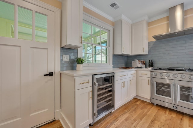 kitchen featuring range with two ovens, wall chimney exhaust hood, light wood-type flooring, white cabinetry, and beverage cooler