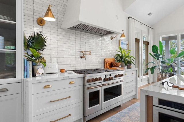 kitchen with range with two ovens, tasteful backsplash, custom range hood, and white cabinets