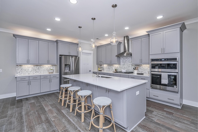 kitchen with a kitchen island with sink, wall chimney range hood, hanging light fixtures, appliances with stainless steel finishes, and a breakfast bar area