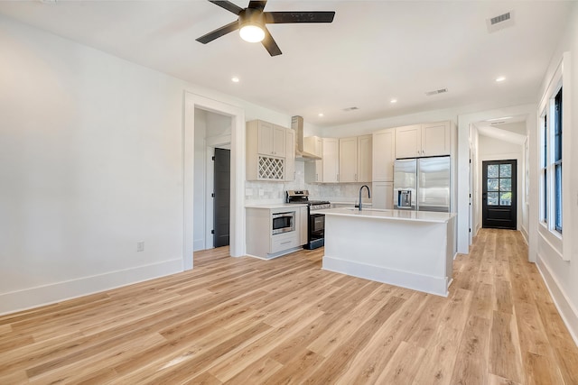 kitchen with wall chimney range hood, stainless steel appliances, a kitchen island with sink, and light hardwood / wood-style flooring