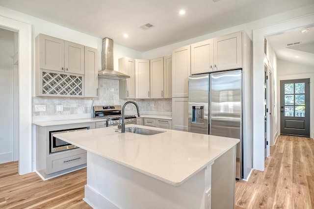 kitchen with light stone countertops, appliances with stainless steel finishes, sink, wall chimney range hood, and a center island with sink