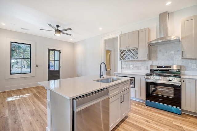 kitchen featuring appliances with stainless steel finishes, wall chimney exhaust hood, a kitchen island with sink, sink, and light hardwood / wood-style flooring