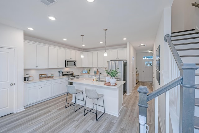 kitchen with white cabinetry, an island with sink, stainless steel appliances, decorative light fixtures, and light hardwood / wood-style flooring