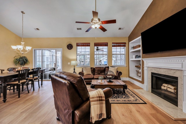 living room with ceiling fan with notable chandelier, built in shelves, a fireplace, vaulted ceiling, and light hardwood / wood-style flooring