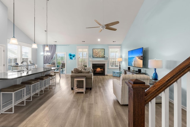 living room featuring ceiling fan, high vaulted ceiling, light wood-style flooring, a fireplace with flush hearth, and stairs