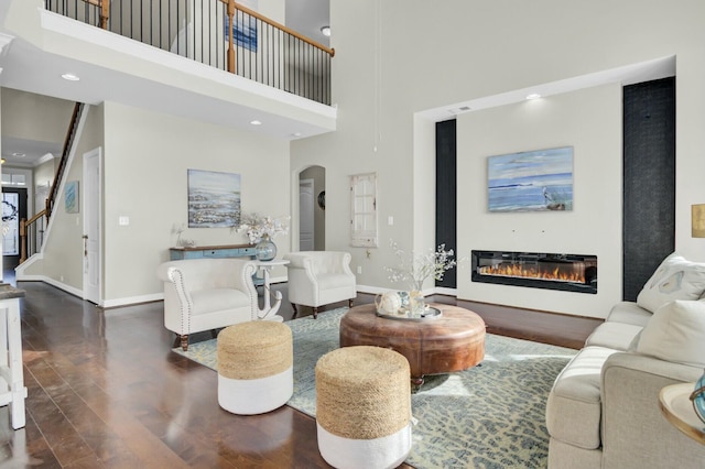 living room with arched walkways, a towering ceiling, a glass covered fireplace, wood finished floors, and baseboards