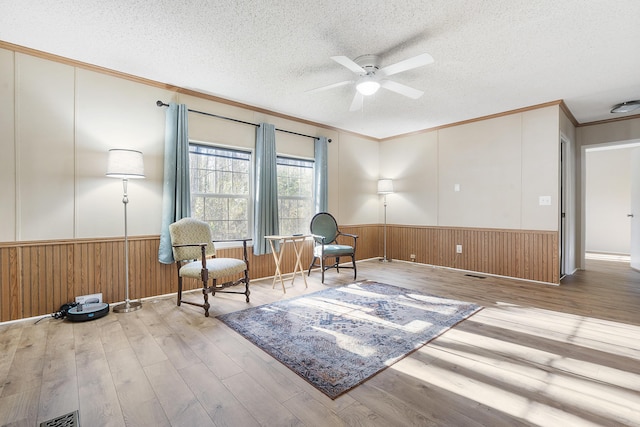 sitting room with a textured ceiling, crown molding, a ceiling fan, and wainscoting
