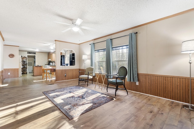 living area with a textured ceiling, a ceiling fan, a wainscoted wall, and crown molding