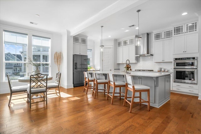 kitchen with a kitchen island with sink, wood-type flooring, hanging light fixtures, wall chimney exhaust hood, and white cabinetry