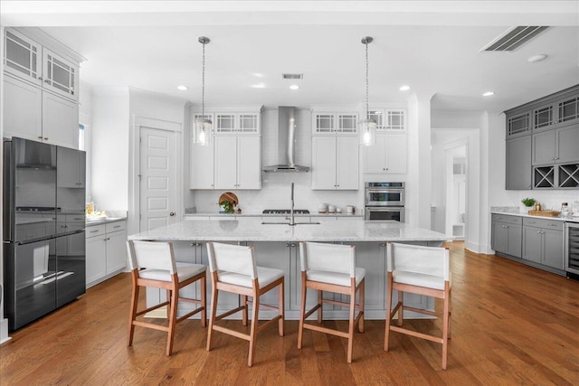 kitchen with wall chimney range hood, backsplash, stainless steel appliances, gray cabinetry, and a kitchen island with sink