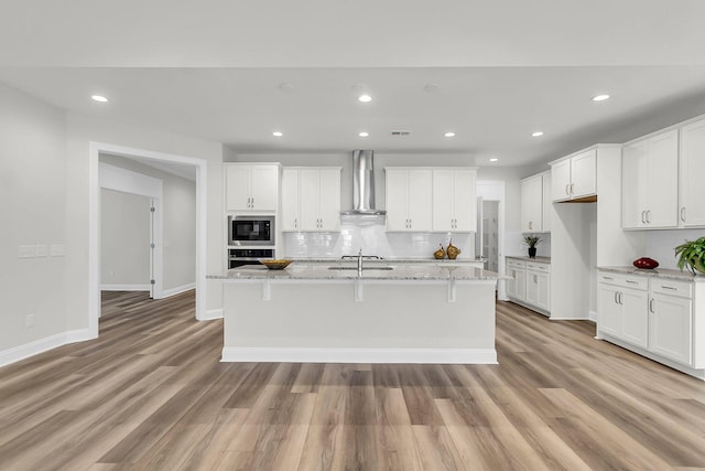 kitchen featuring built in microwave, white cabinets, an island with sink, and wall chimney range hood