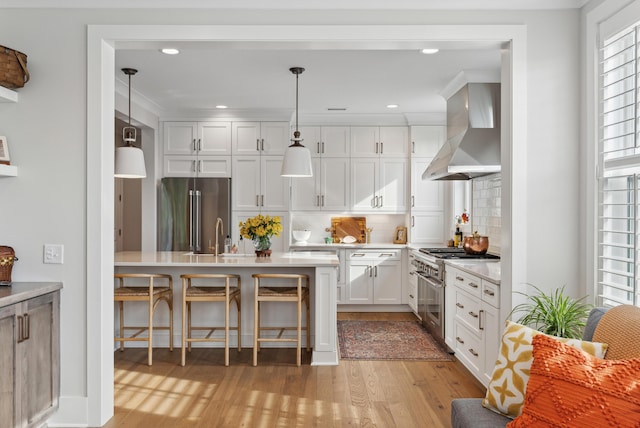 kitchen featuring wall chimney range hood, high end appliances, light countertops, light wood-style floors, and a breakfast bar
