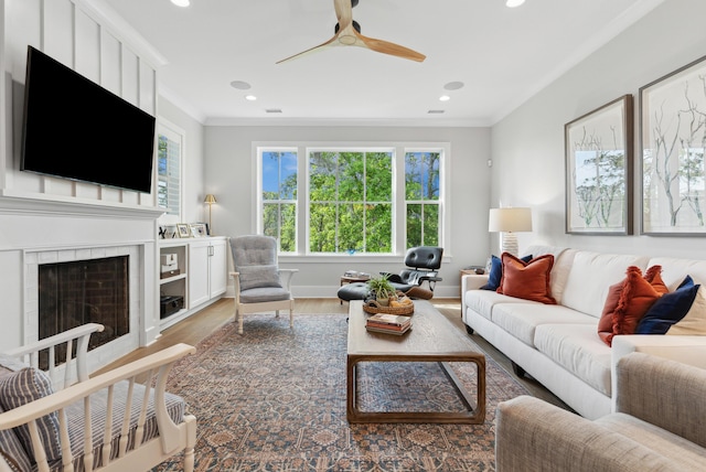 living area featuring crown molding, light wood finished floors, a tiled fireplace, a ceiling fan, and recessed lighting
