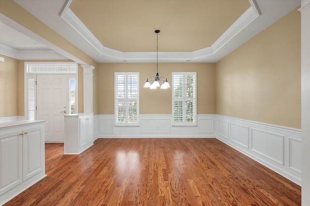 unfurnished dining area featuring crown molding, decorative columns, a notable chandelier, a tray ceiling, and wood-type flooring