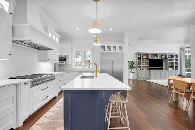 kitchen featuring premium range hood, decorative light fixtures, sink, built in appliances, and a center island with sink