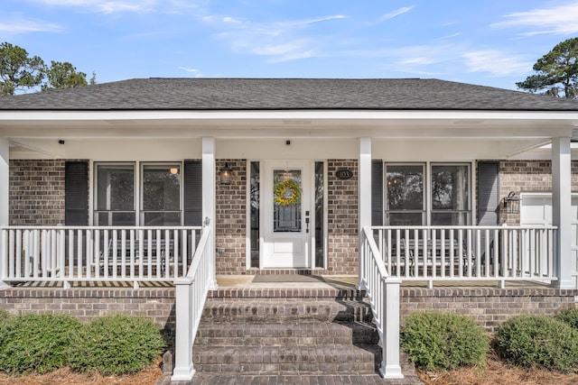 view of exterior entry featuring covered porch, a shingled roof, and brick siding