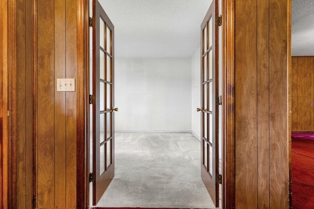 corridor featuring french doors, wood walls, light colored carpet, and a textured ceiling