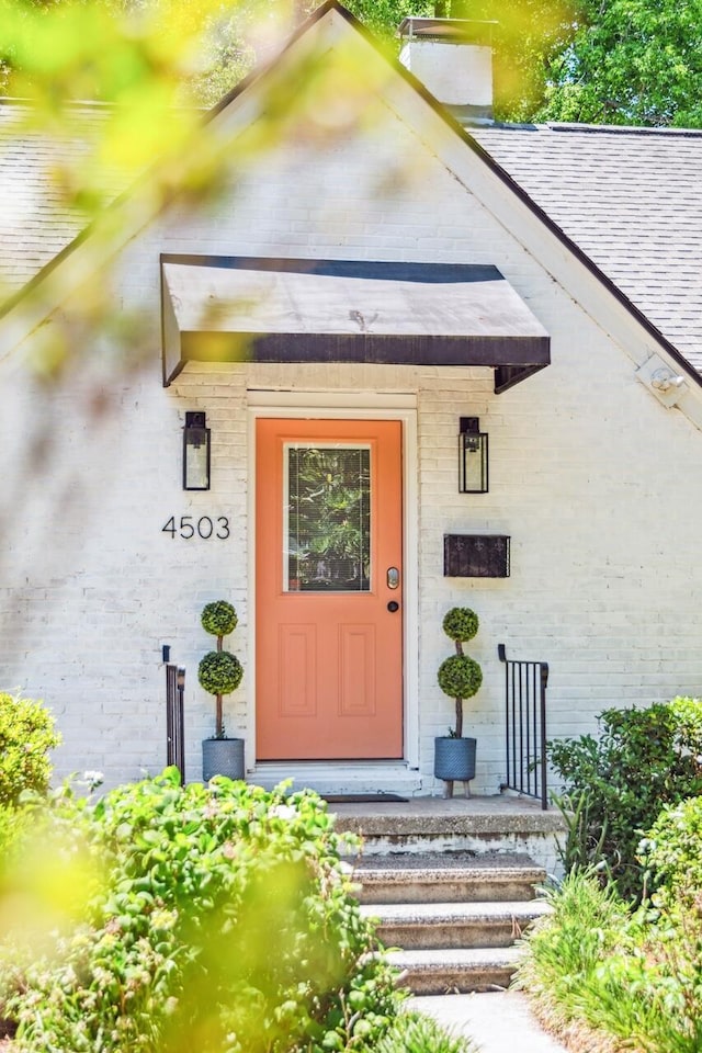 doorway to property with brick siding, roof with shingles, and a chimney