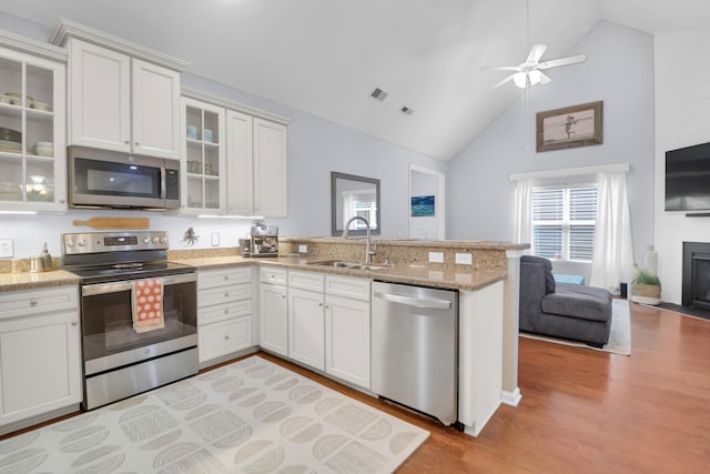 kitchen featuring stainless steel appliances, a sink, a peninsula, light wood finished floors, and high vaulted ceiling