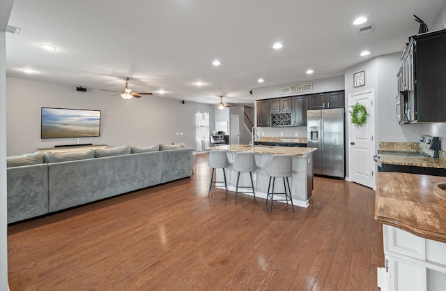 kitchen with appliances with stainless steel finishes, a breakfast bar, light stone countertops, dark wood-type flooring, and a center island with sink