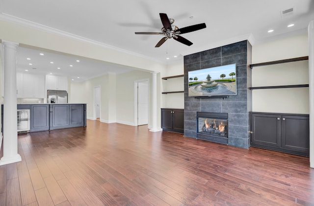 unfurnished living room with hardwood / wood-style flooring, a tiled fireplace, crown molding, ceiling fan, and decorative columns