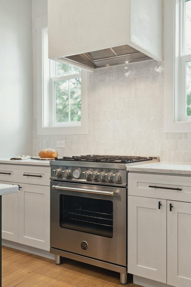 kitchen featuring exhaust hood, white cabinets, high end stainless steel range oven, decorative backsplash, and light hardwood / wood-style floors