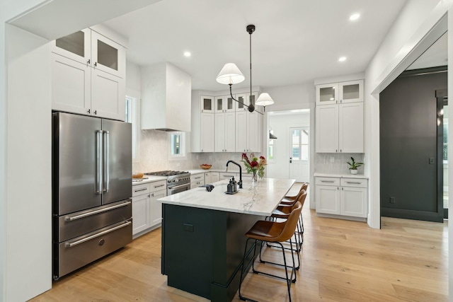 kitchen featuring light stone countertops, custom exhaust hood, high end appliances, a kitchen island with sink, and white cabinetry