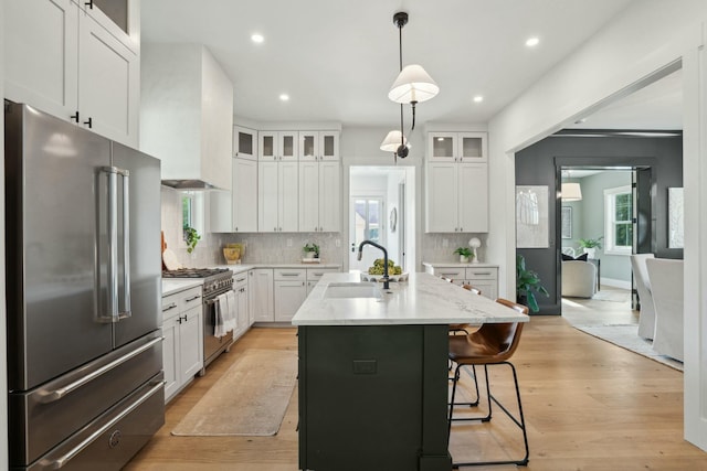 kitchen featuring high quality appliances, premium range hood, white cabinetry, a center island with sink, and a sink
