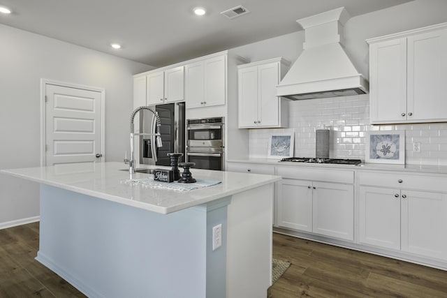 kitchen with appliances with stainless steel finishes, a kitchen island with sink, custom range hood, and white cabinets