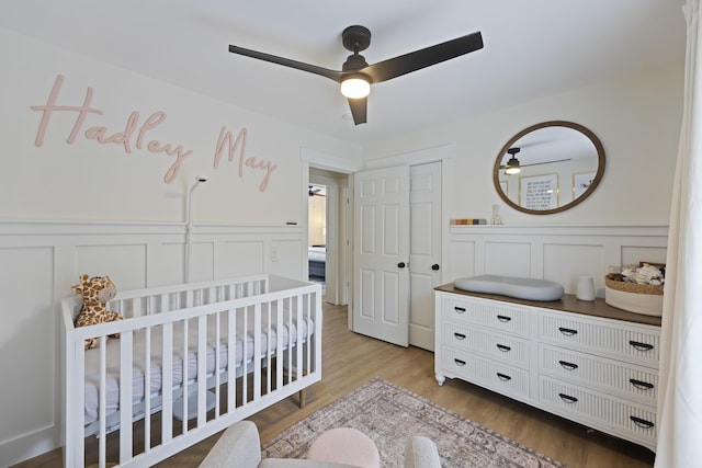 bedroom featuring light wood finished floors, a wainscoted wall, a ceiling fan, and a nursery area