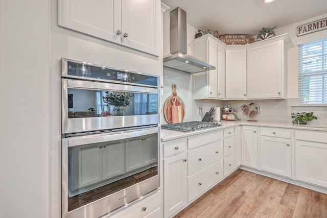 kitchen with stainless steel appliances, wall chimney exhaust hood, light countertops, white cabinets, and backsplash
