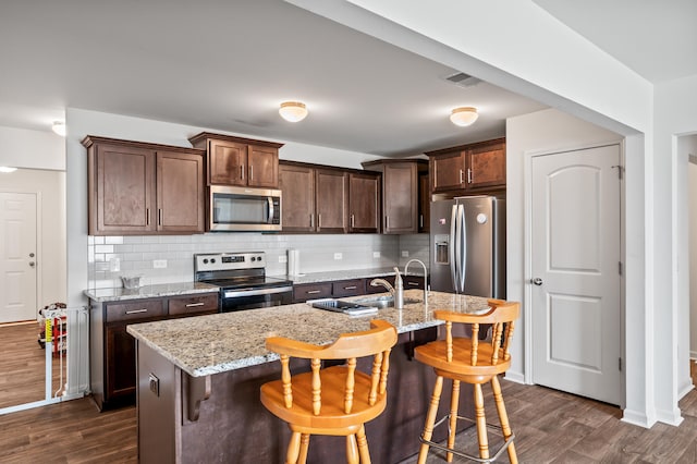 kitchen with appliances with stainless steel finishes, light stone counters, dark hardwood / wood-style floors, a breakfast bar area, and a kitchen island with sink