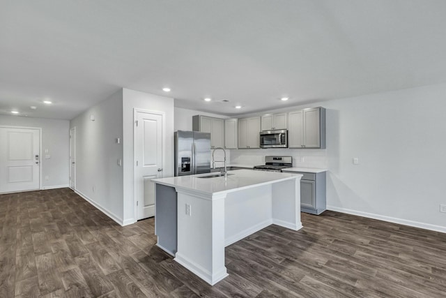 kitchen with a sink, gray cabinetry, an island with sink, appliances with stainless steel finishes, and dark wood-style flooring