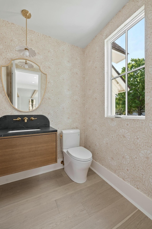 half bath featuring toilet, wallpapered walls, vanity, wood finished floors, and baseboards