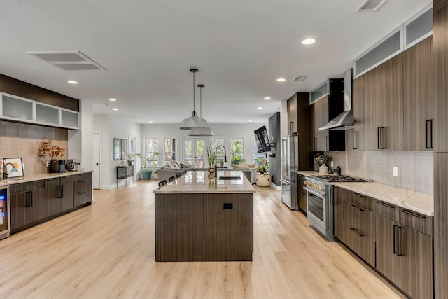 kitchen featuring sink, a kitchen island with sink, hanging light fixtures, stainless steel appliances, and wall chimney exhaust hood