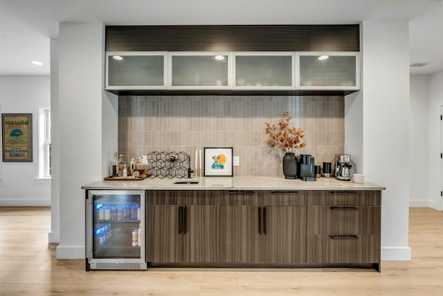 bar with dark brown cabinetry, beverage cooler, light hardwood / wood-style flooring, and backsplash