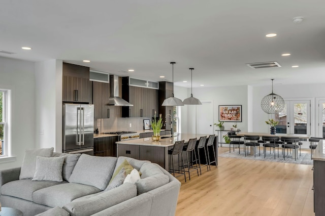 kitchen featuring wall chimney exhaust hood, dark brown cabinets, a center island with sink, pendant lighting, and stainless steel appliances