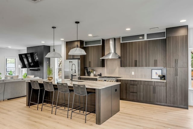 kitchen featuring dark brown cabinetry, sink, hanging light fixtures, a kitchen island with sink, and wall chimney range hood