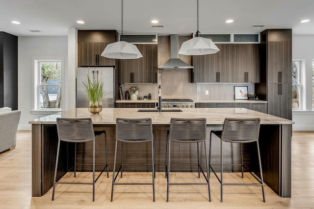 kitchen featuring hanging light fixtures, an island with sink, stainless steel refrigerator, and wall chimney exhaust hood