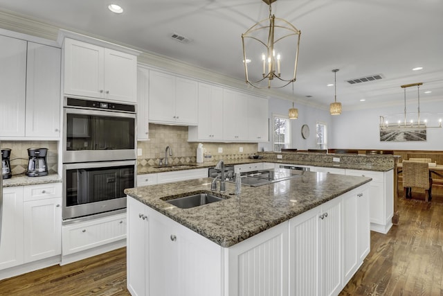 kitchen with white cabinets, stainless steel double oven, hanging light fixtures, and an island with sink