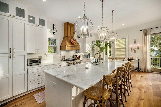 kitchen with custom exhaust hood, light stone counters, a center island with sink, pendant lighting, and white cabinets