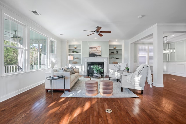 living room featuring built in shelves, ceiling fan with notable chandelier, crown molding, and dark wood-type flooring