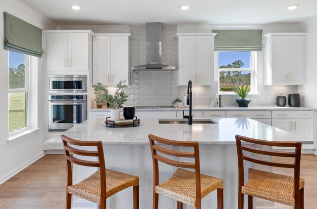 kitchen with appliances with stainless steel finishes, a wealth of natural light, tasteful backsplash, light wood-type flooring, and wall chimney range hood