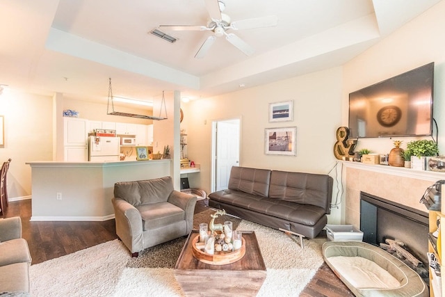 living room featuring dark hardwood / wood-style floors, ceiling fan, a raised ceiling, and a tiled fireplace