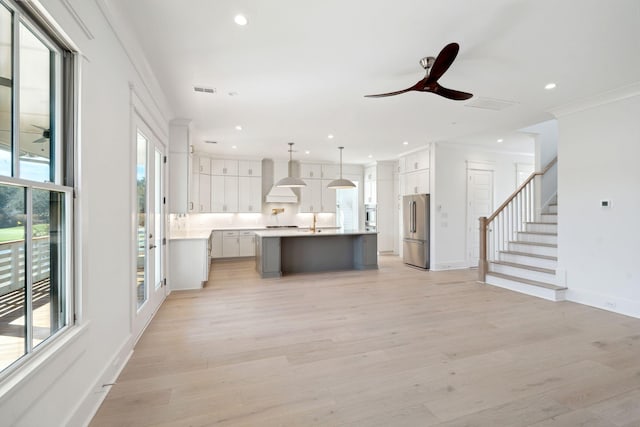 kitchen with high end fridge, white cabinetry, an island with sink, pendant lighting, and wall chimney range hood