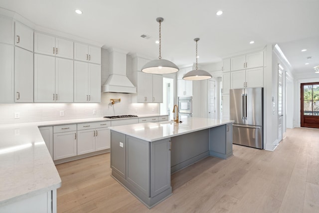 kitchen with high end refrigerator, gas cooktop, custom range hood, a kitchen island with sink, and white cabinets